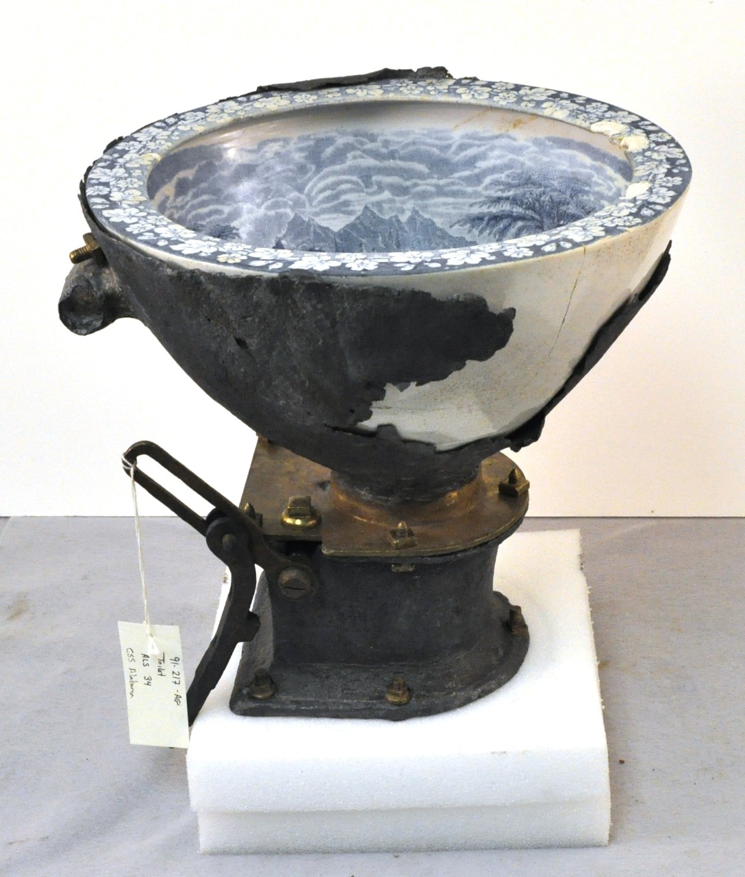 <p>A flushing toilet recovered from CSS Alabama. A large porcelain bowl decorated with boating and country scenes set in a lead base that tapers down onto a large square brass washer. A circular hole is made at the bottom of the bowl where a round brass door swings open and shut. Connected to the brass washer is brass handle which would allow you to operate the brass door. The brass washer is screwed into another square lead piece below it and abruptly tapers to a circular lead piece.</p>
