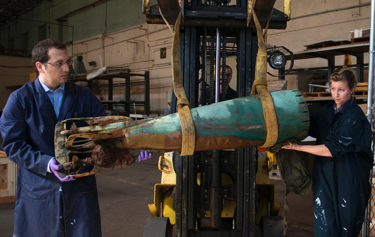 Two archaeologist from the NHHC Underwater Archaeology Branch stand on either side of the tail section of the Howell Torpedo to keep it balanced as the forklift carries it to its holding tank. 