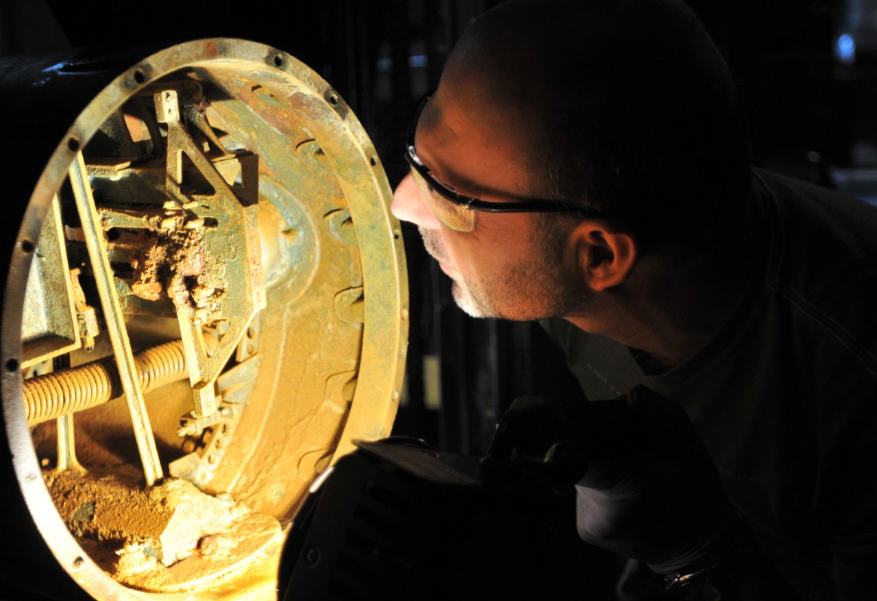 Conservator Paul Mardikian uses a flash light to view the interior of the tail end of the Howell Torpedo to assess the corrosion and state of preservation. 