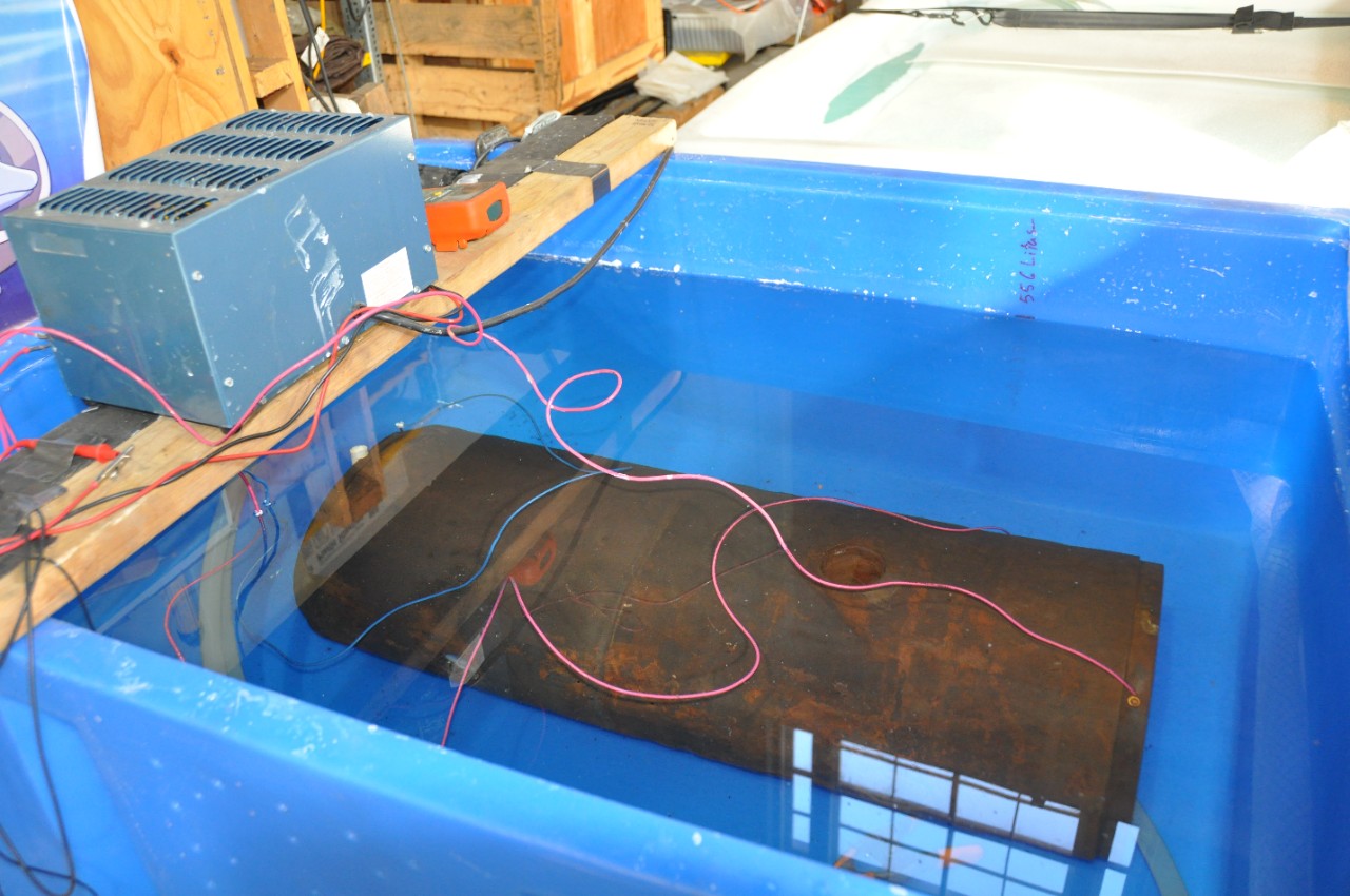 <p>The mid-section of Howell torpedo undergoing electrolytic reduction while submerged in a chemical solution in a blue plastic treatment tank. Wires project from the direct circuit power supply set on a wood plank above the tank and connected to the exterior and interior of the mid-section.</p>
