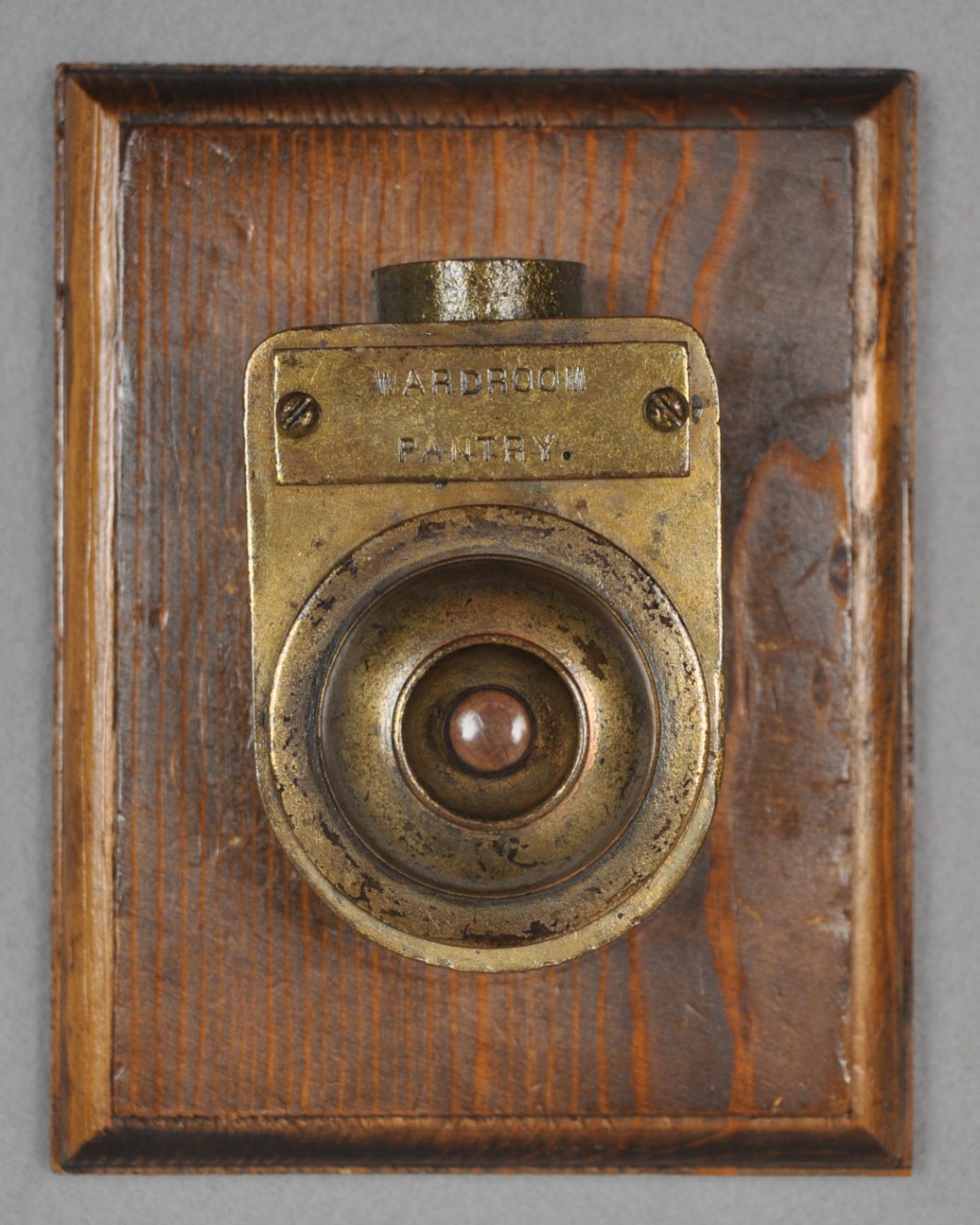 <p>A small brass box with a raised circular button in the middle. Above the circular button is a brass tag with the words “Wardroom Pantry” stamped into it. At the top of the brass box is a small circular tube for cable to run through. It is mounted on a square wooden board.</p>
