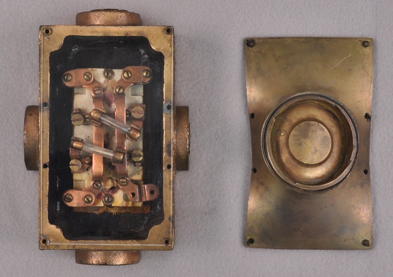 <p>A rectangular brass box with a crushed detached faceplate. Inside the box is a small rectangular ceramic block with copper strips and screws attached to it. Within the copper strips is two glass tubes called fuses. On each side of the box are raised circular tubes. Located on the right side of the box, is the detached faceplate with eight screw holes.</p>
