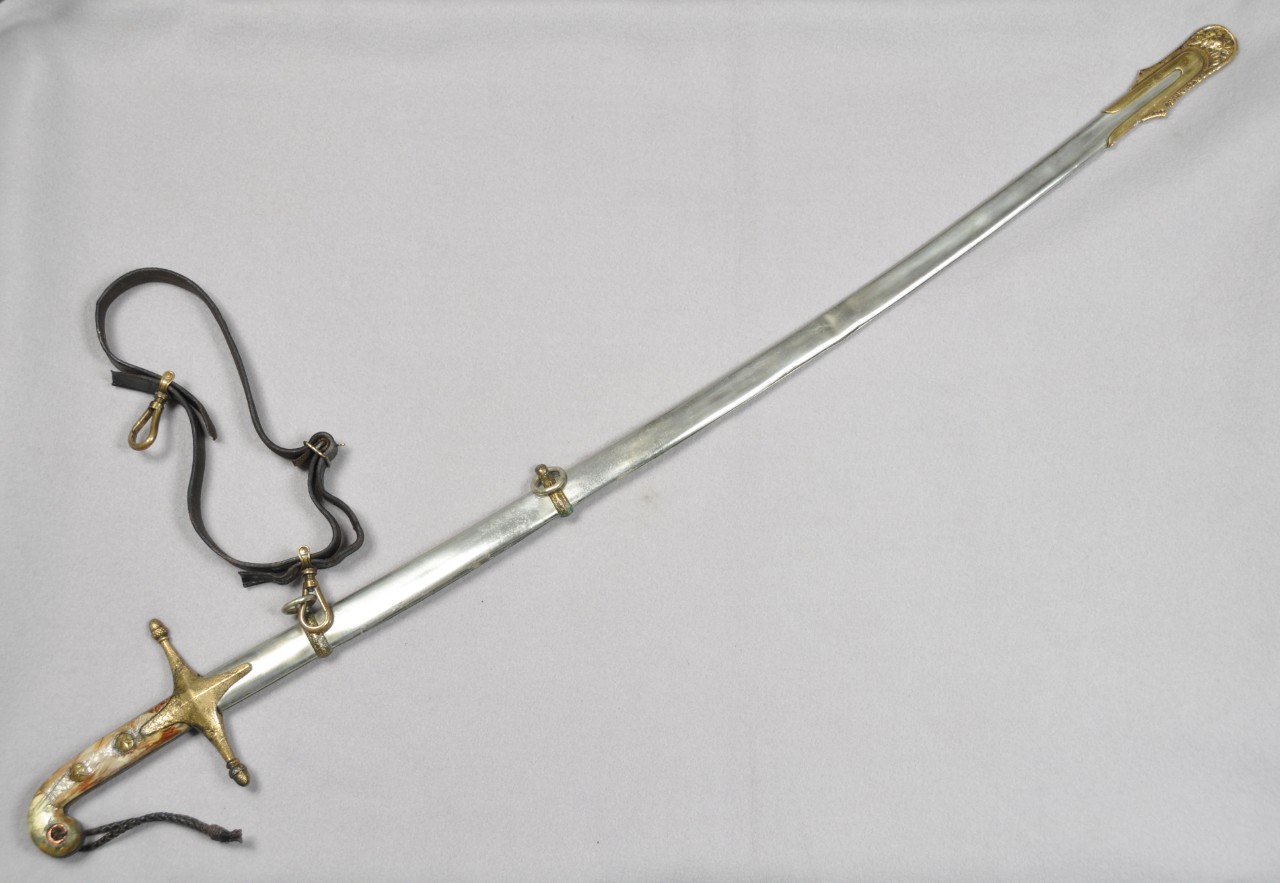 <p>A long sword with wooden grip. The grip has a hole at the bottom with a braided leather strip. In the middle of the grip are two brass stars. Above the grip is a brass cross-guard with knobs at each end. The sword blade is sheath with a silver colored scabbard. The scabbard has two brass fittings located close the grip with rings attached. A bent leather strip is attached to one of these rings with a brass clip. The other clip is located further up the leather strap. At the tip of the scabbard is a decorated brass chape or tip.</p>
