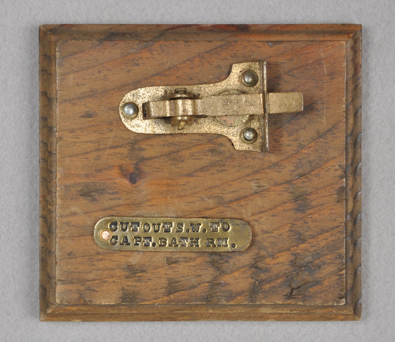 <p>Attached to a square wooden piece of wood is a brass cabinet latch and below the latch is an oblong brass tag with the woods “Cut Out S.W. To Capt. Bath Rm.” stamped on it.</p>