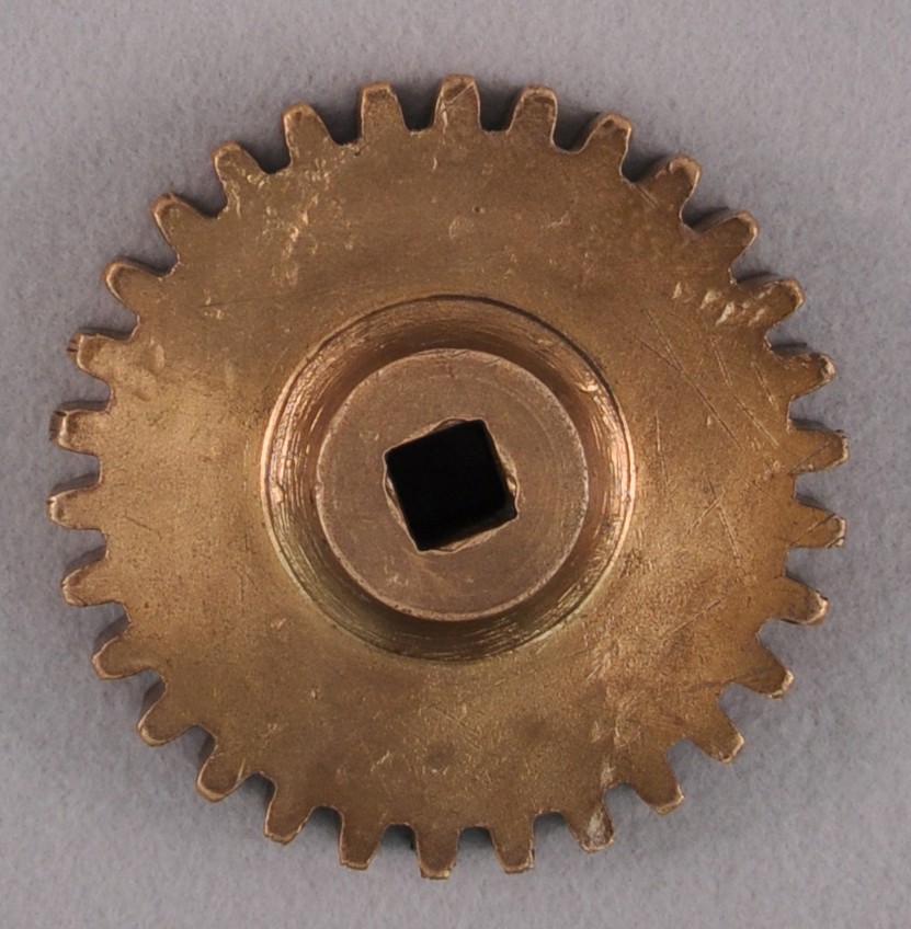 <p>A circular brass gear with small prongs along the outside. In the middle is a raised brass circle with a square hole cut in the middle to place a square object inside.</p>