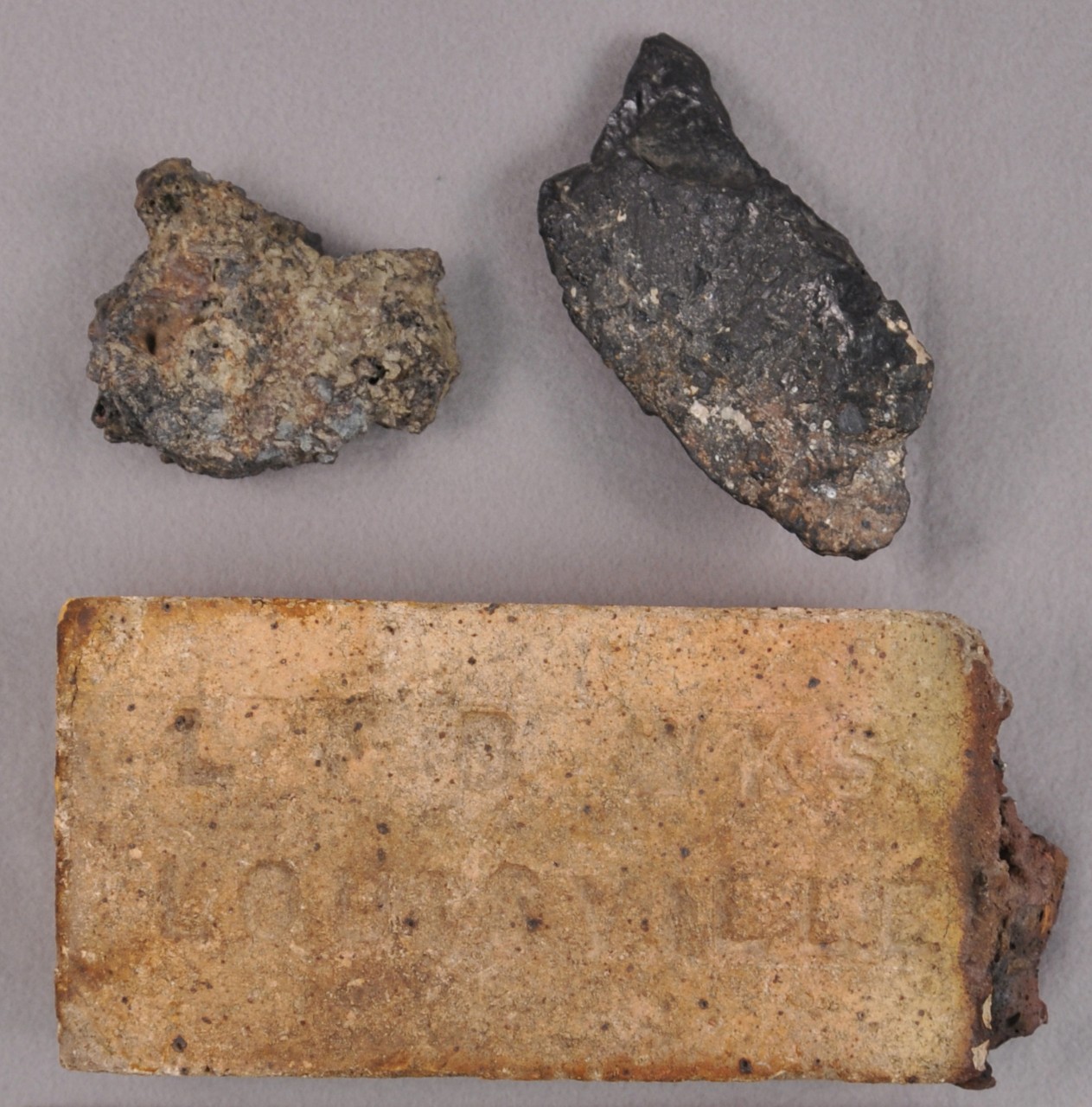 <p>At the top are two irregular shaped pieces of coal. Below the coal is a brick with the words “LFB WKS/LOUISVILLE” on the side that is shown in the picture. On the right corner of the brick is a piece of coal slag attached.</p>
