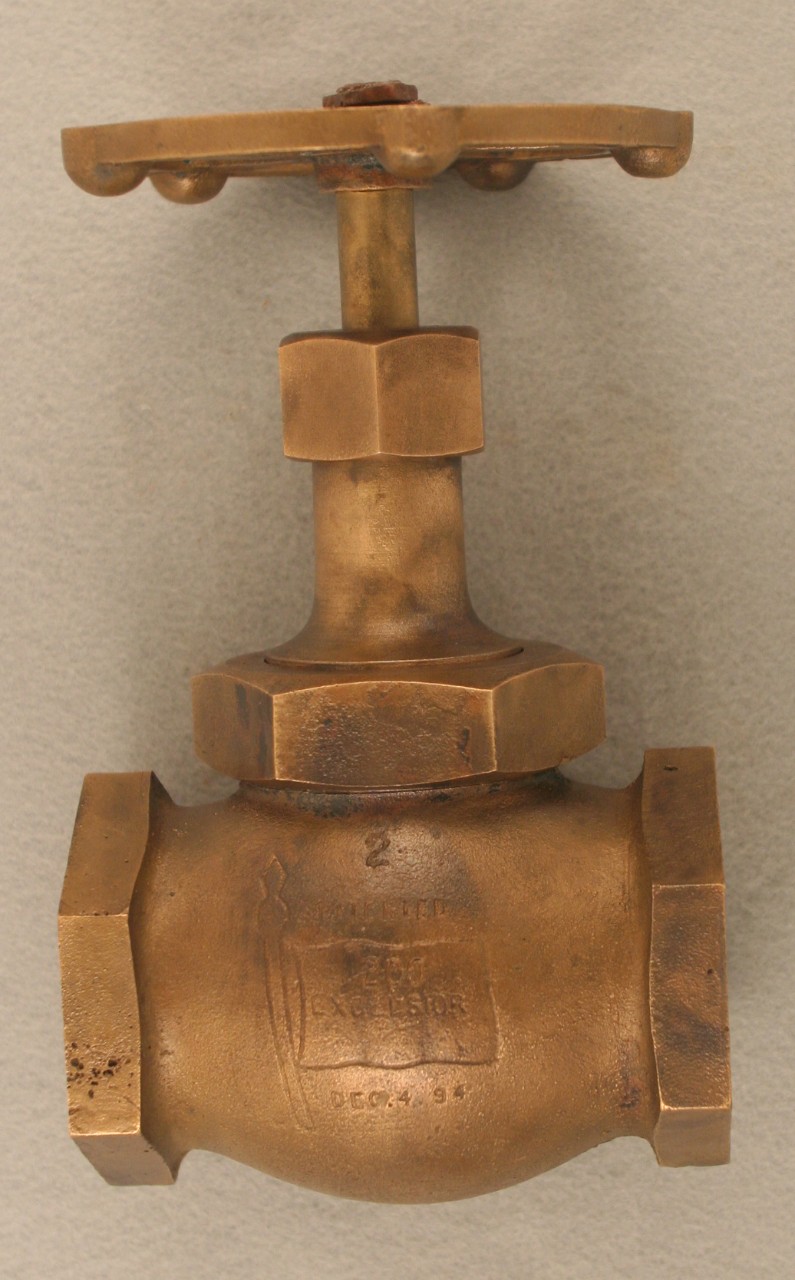 <p>A brass steam valve with a circular valve wheel at the top connected to a horizontal tube with two large nuts. The last large nut is screwed into a vertical fitting with two openings at each end. In the middle of fitting is the number “2” with the word “Patented” below followed by an etching of a flag pole and flag. Inside the flag is the letters “200 / Excelesior”. Below the flag is the date “Dec. 4 94”.</p>