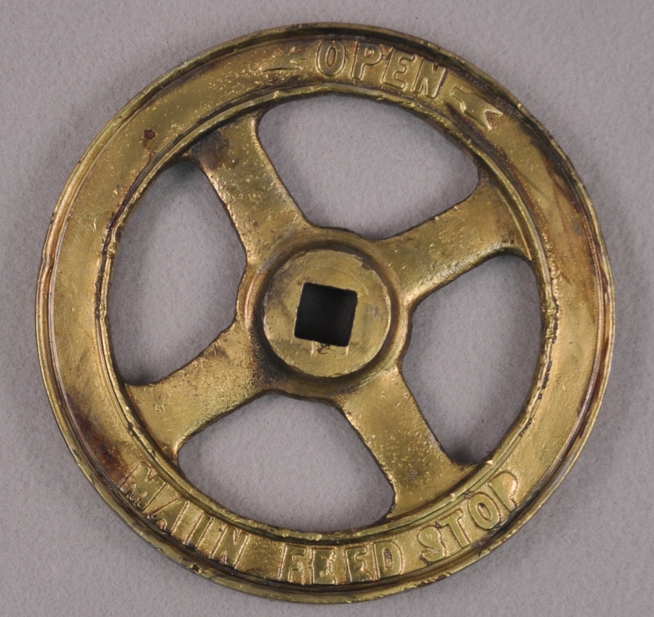 <p>A brass valve wheel with a raised circle in the middle. A square hole is cut in the middle of this circle. At the top of the valve are the words “OPEN” with an arrow point left and the words at the bottom are the words “Main Feed Stop”.</p>