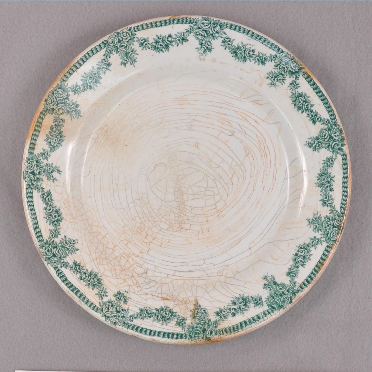 <p>A white stoneware plate recovered from USS San Diego with the glazed cracked. It has a continuous green flower and vine pattern around the border.</p>