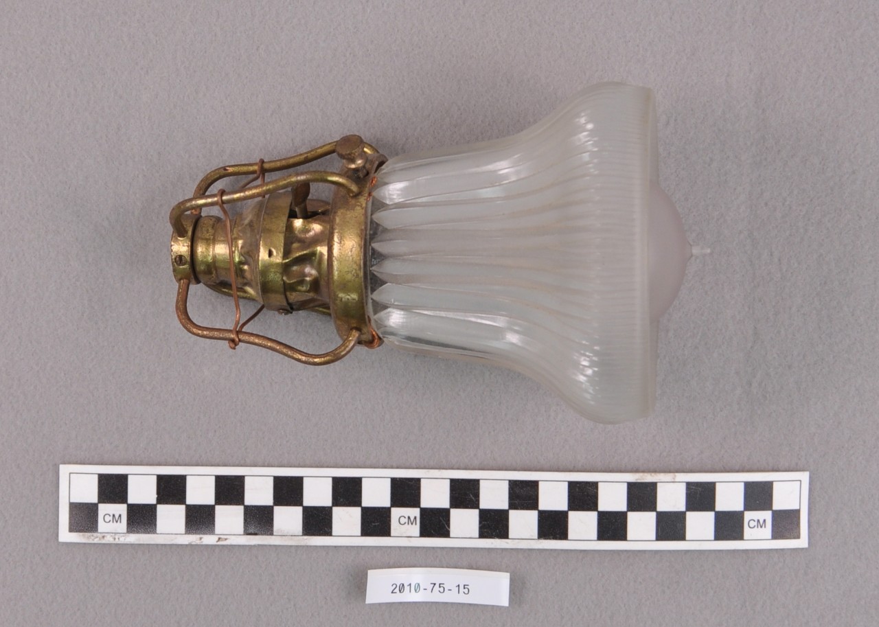 <p>At the top is a brass light bulb holder with four bent rods connecting the top of the holder to the bottom. On the left side of the holder is a light switch. Screwed into to the brass holder is a decorated glass fixture and a historic light bulb.</p>

