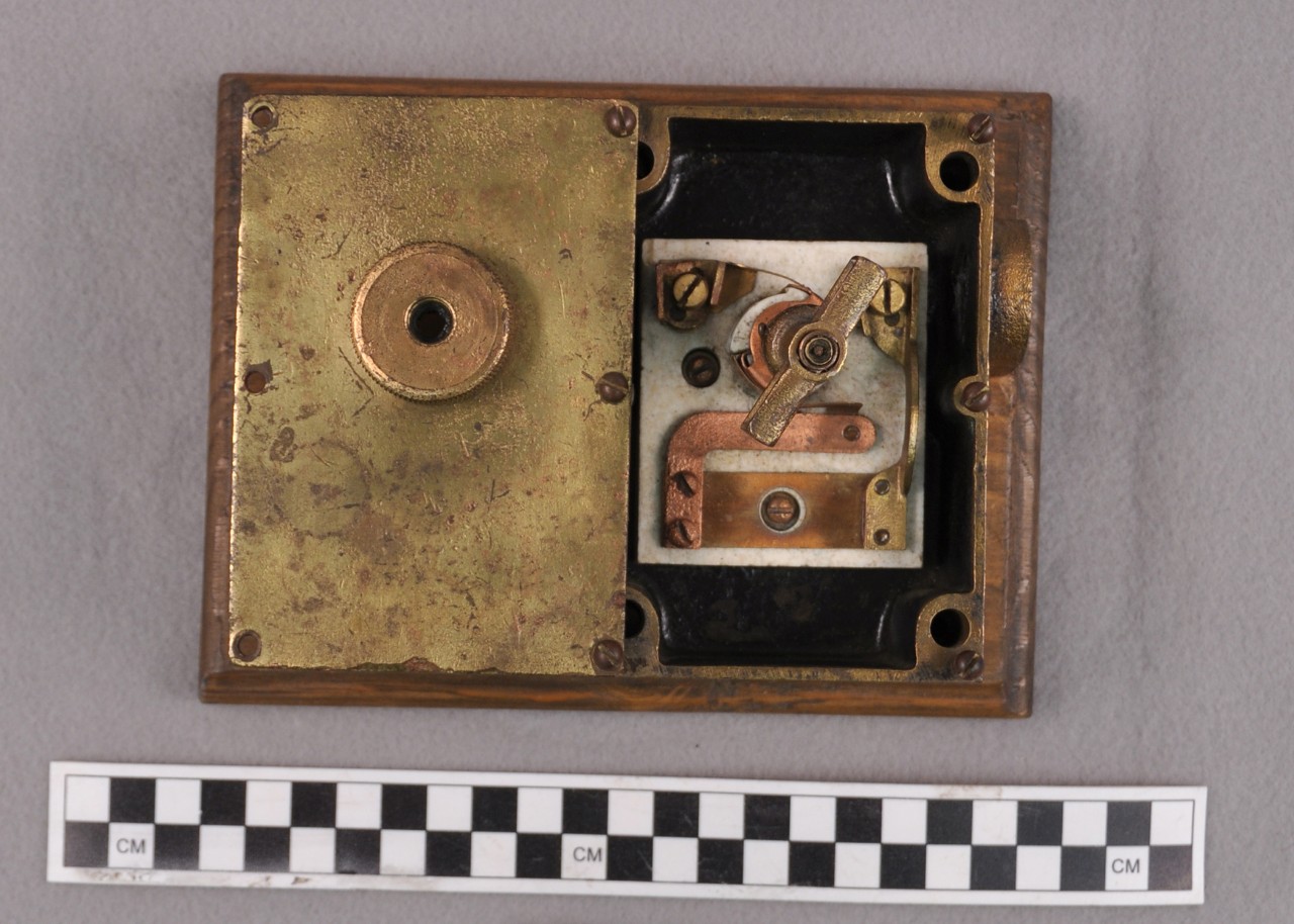 <p>A small rectangular brass box with the faceplate opened and attached to the left of the box. The faceplate has three screw holes on the left and three screws attached to the box on the right. In the middle is a small circular knob for the switch. The box contains a small square ceramic block inside with copper and brass fittings. On the right side of the box is a small circular tube for electrical wires to go through.</p>
