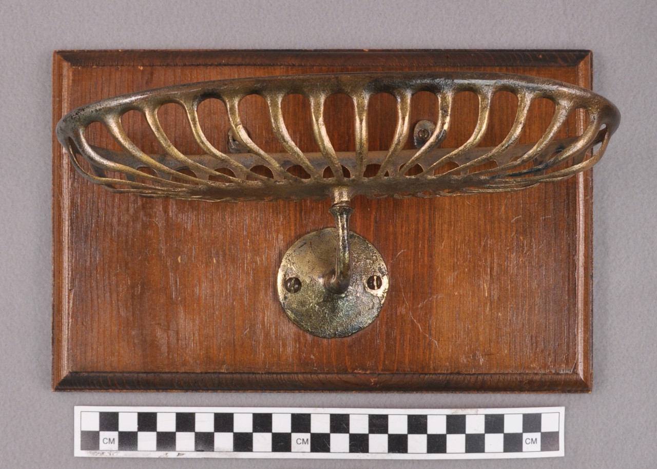 <p>An oblong brass soap holder. The rim is solid with individual rods attached around the rim and solid base leaving holes between the rods. The solid base is attached to a curved brass rod that is attached to a circular disk that is mounted to a square wooden block.</p>
