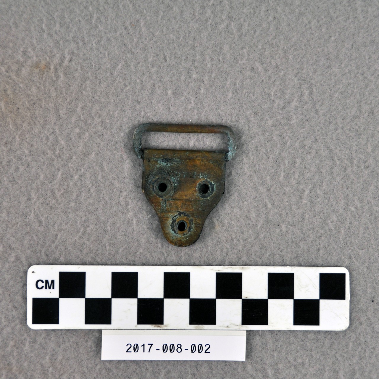 <p>A small brass strap adjuster for a sling, shaped like an upside-down pentagon with three holes through it. There is text on the face that reads “PATD 7-21-14 NOBUCKL REG… PAT OFF”</p>