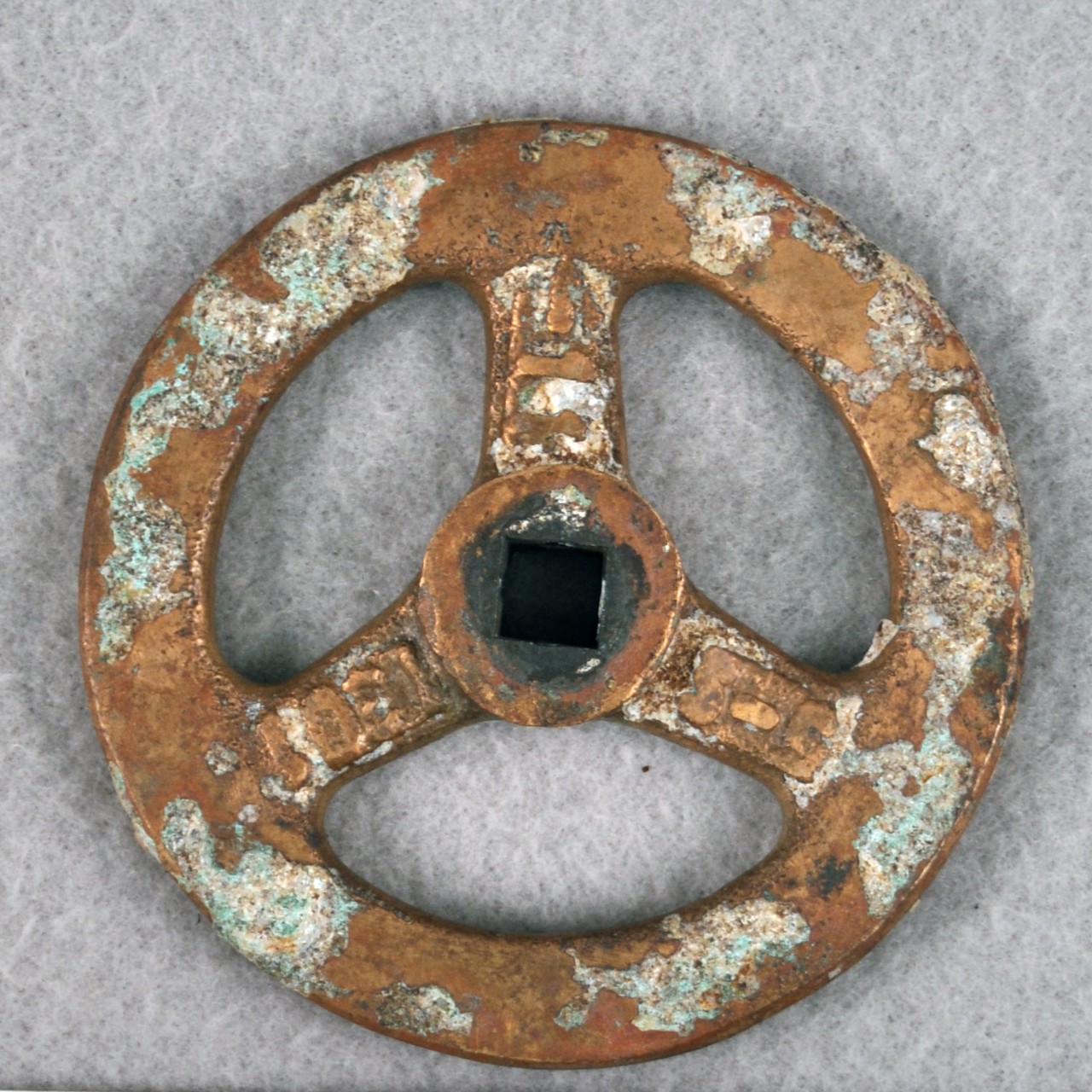 <p>A small brass wheel belonging to a valve. There are three metal spokes on the face, on which there are raised numbers. The numbers are partially obscured by white corrosion; they read “1301”, “…11”, “5-5”.</p>