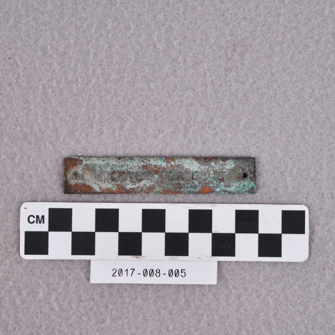 <p>A small, rectangular strip of brass covered with white and green corrosion. There is a hole on one end and a rivet on the other. There is text stamped on face that reads “GENERATOR FIELD”.</p>
