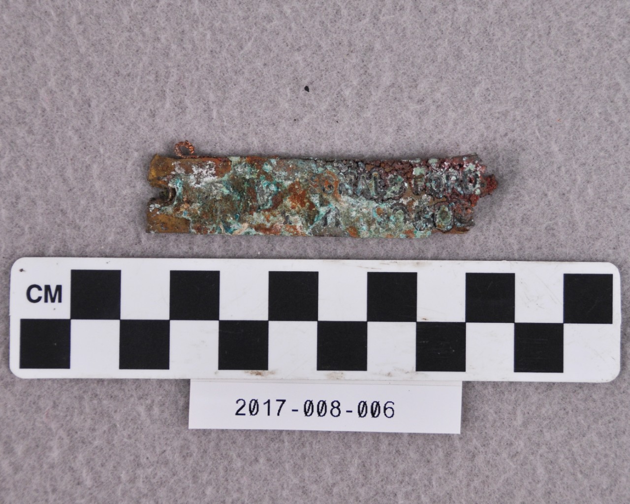<p>A small, rectangular strip of brass covered in white, red, and green corrosion. There is visible writing on the face of it that reads “…GNALS FORD…”.</p>