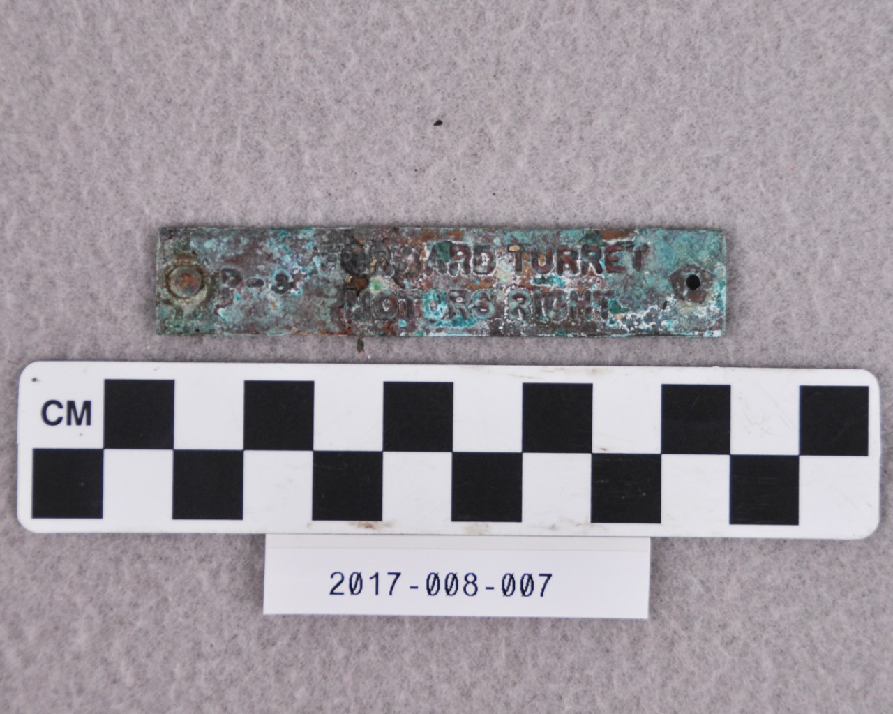 <p>A small, rectangular strip of brass covered in white, green, and red corrosion. There is a hole through one end and a rivet through the other. There is text stamped on the face that reads “P-9 FORWARD TURRET MOTORS RIGHT”.</p>