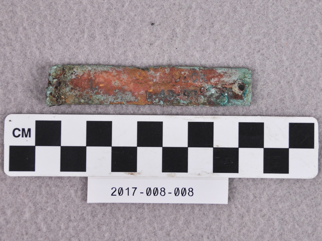 <p>A small, rectangular strip of brass with the edges covered in green and white corrosion. There are two small holes through either end. There is text stamped on the face that reads “…ECK… -43-87. …”.</p>