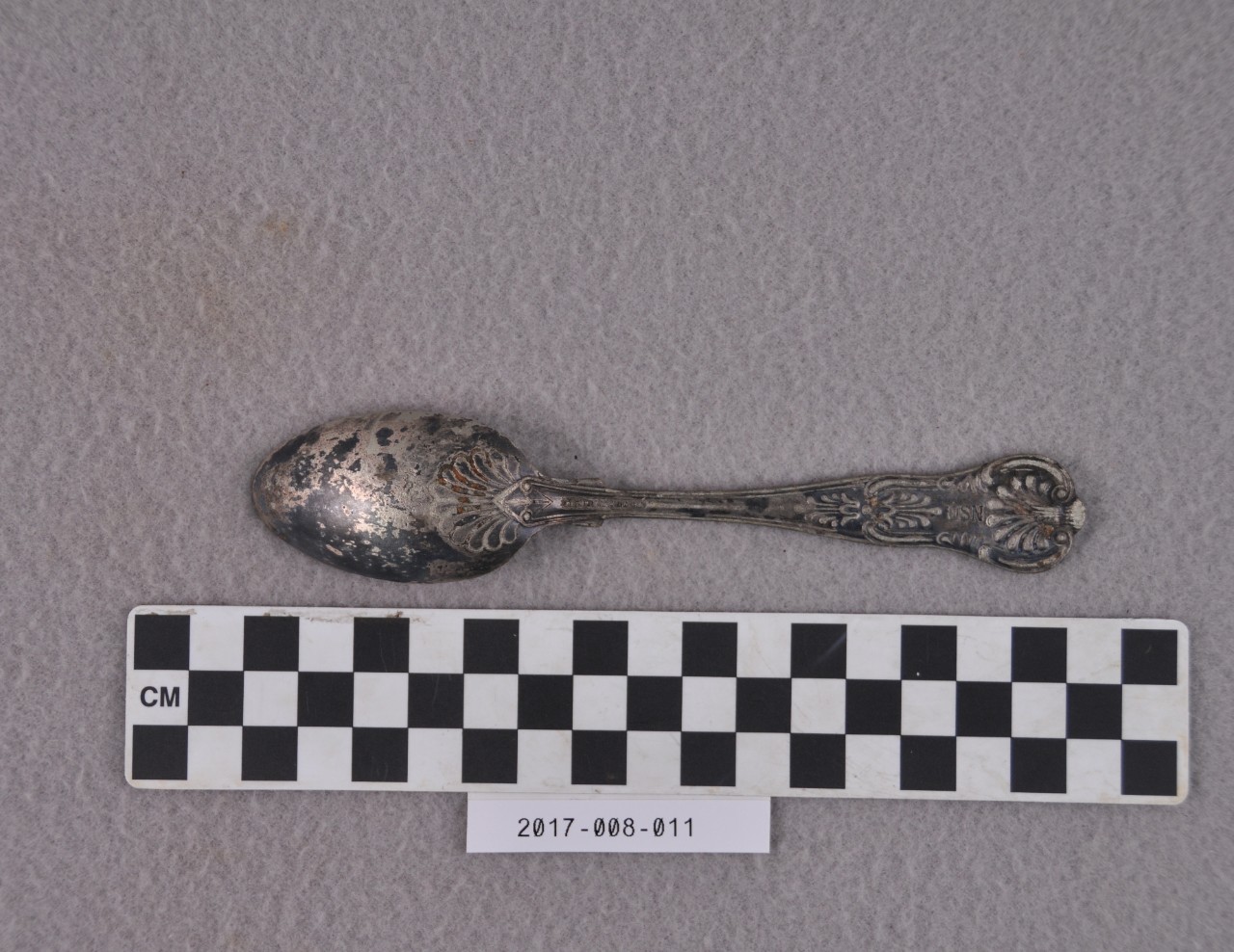 <p>A decorated silver-plated teaspoon. There is partially legible writing stamped on the back of the handle that reads “… R WALLACE” and “USN”.</p>
