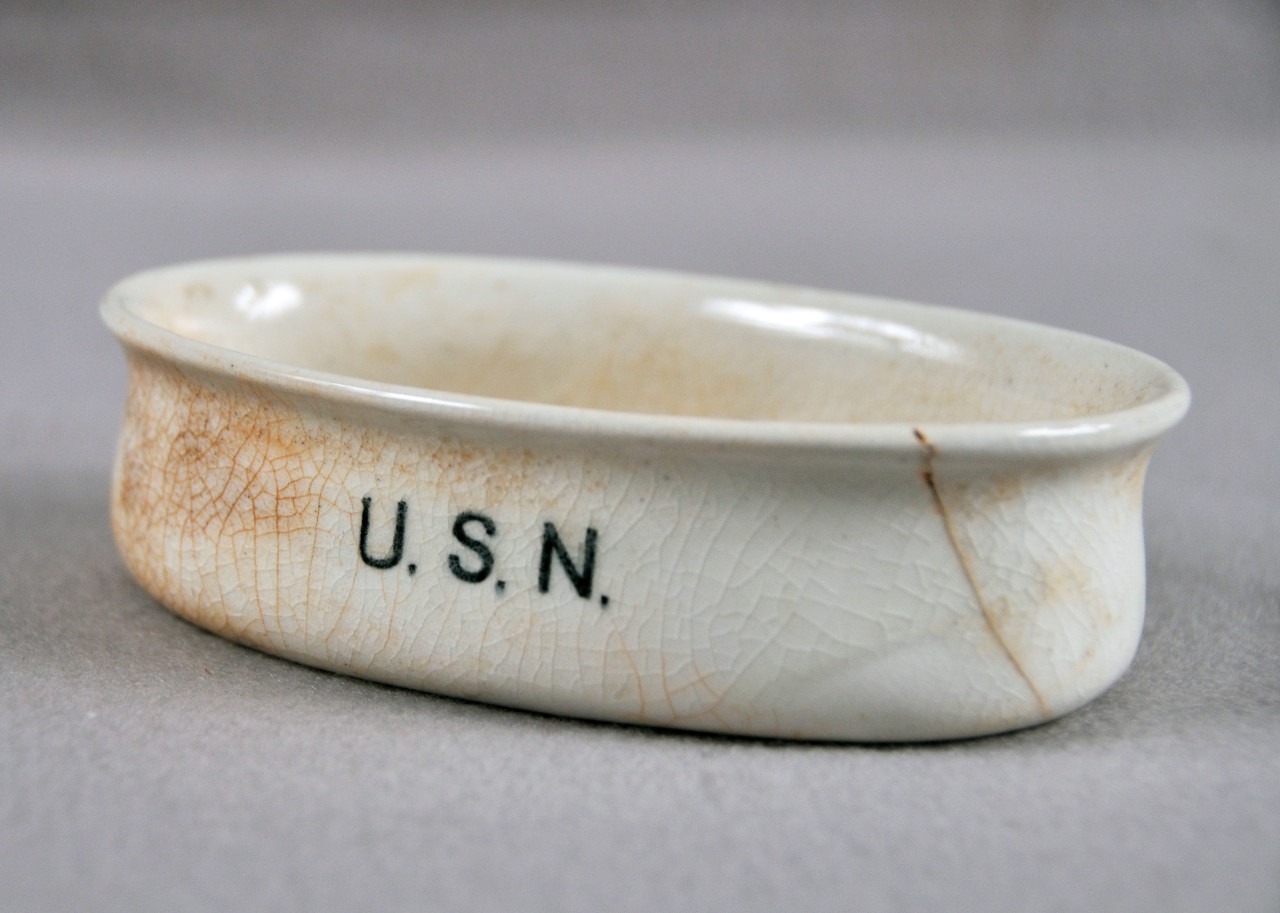 <p>A white ceramic soap dish with the letters “USN” on one side. The surface of the dish is crackled.</p>
