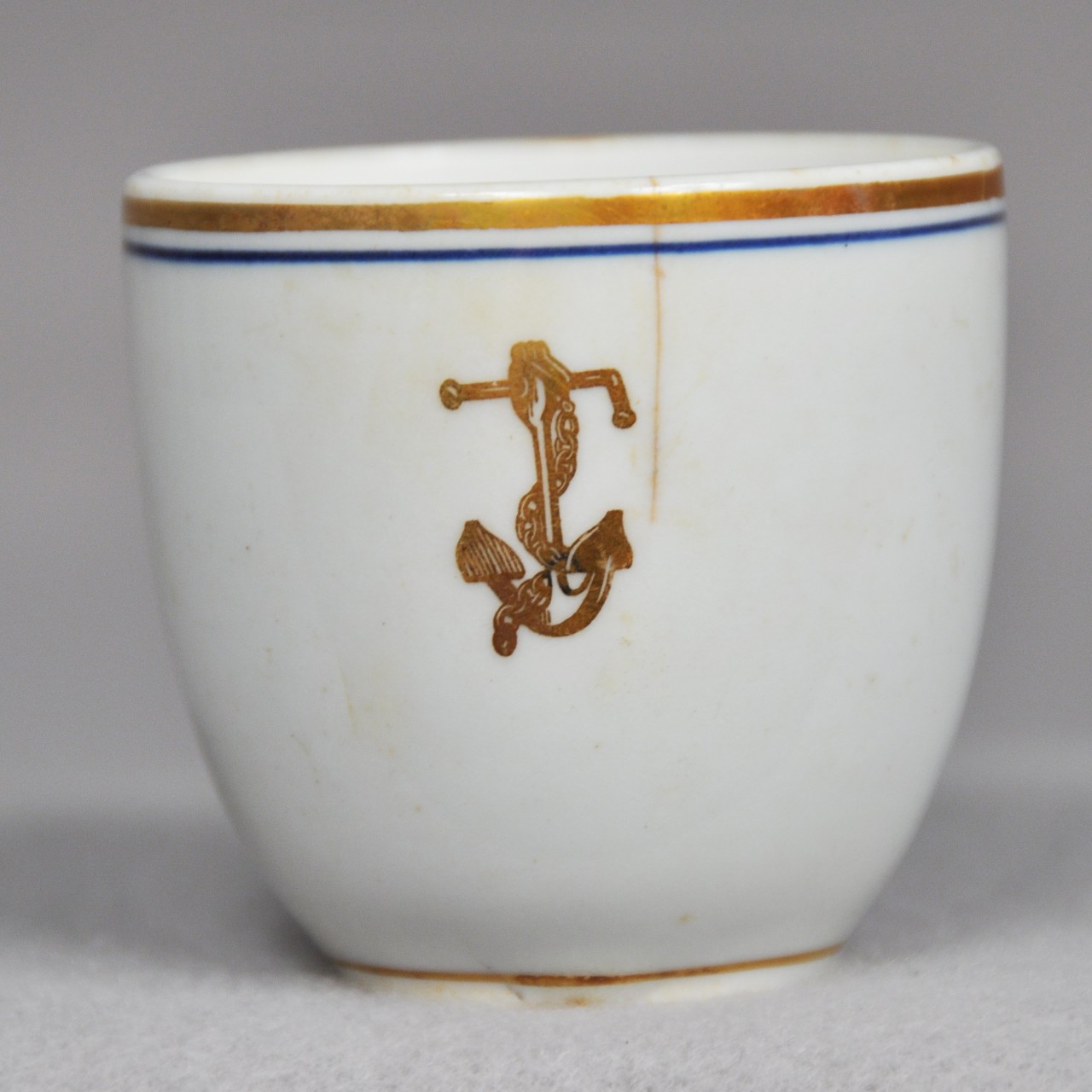 <p>A small ceramic coffee cup. There is a golden ring and a blue ring around the rim of the cup. There is a golden anchor with a chain wrapped around it on the exterior of the cup.</p>
