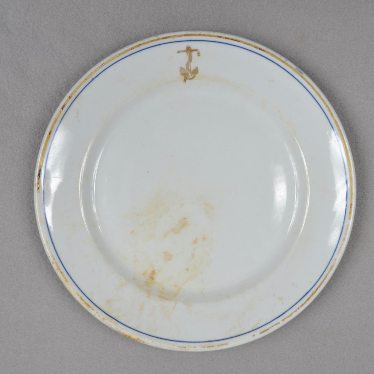 <p>A white ceramic plate with a gold ring and blue ring around the edge. There is also a golden anchor with a chain around it on the lip.</p>