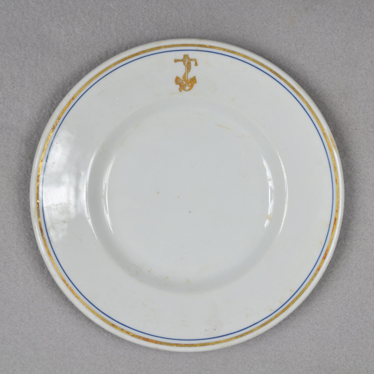 <p>A white ceramic plate with a gold and blue ring around the edge of the face of the plate. There is also a gold anchor with a chain wrapped around it on the lip.</p>