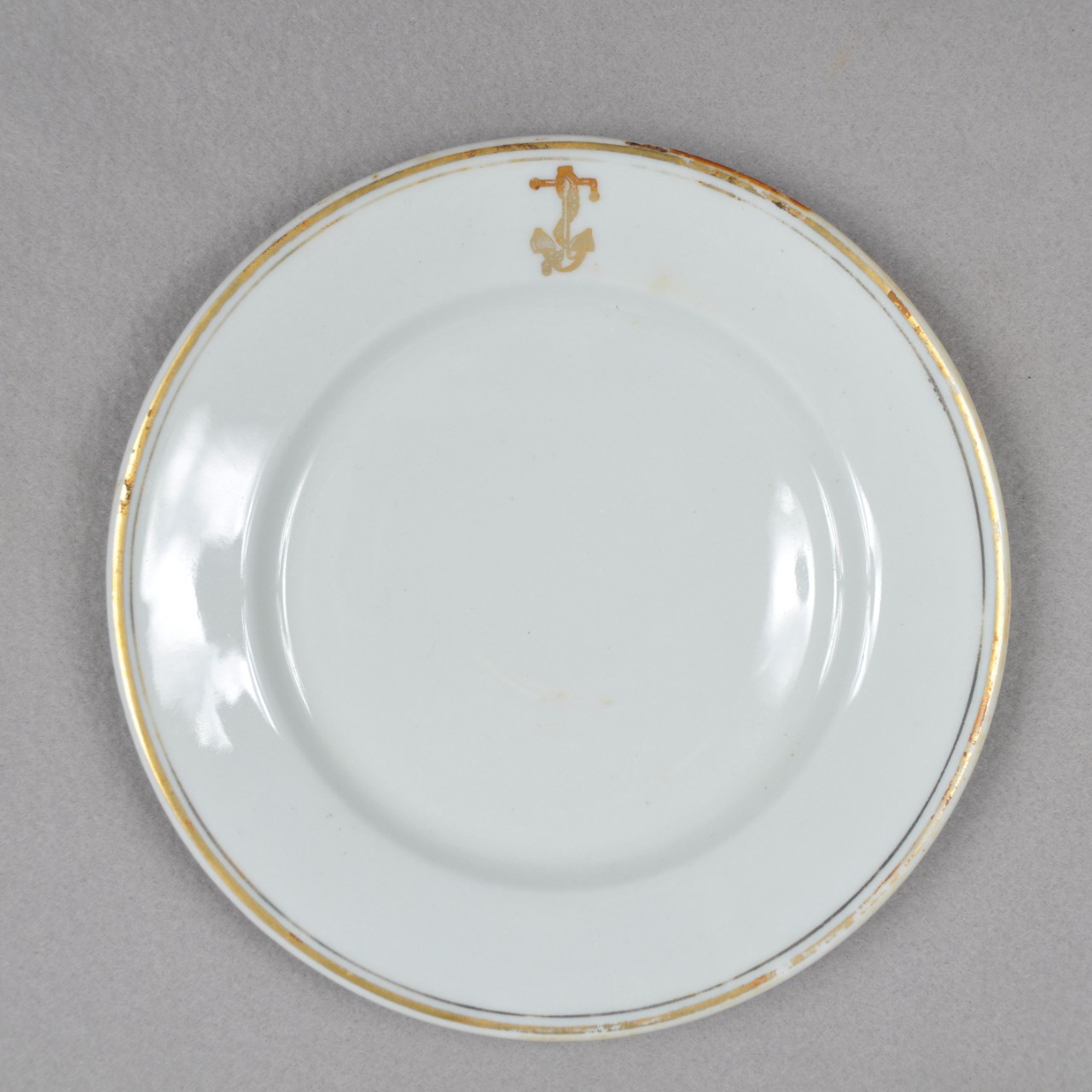 <p>A white ceramic plate with two concentric gold rings around the edge on the face of the plate. There is also a gold anchor with a chain wrapped around it on the lip.</p>
