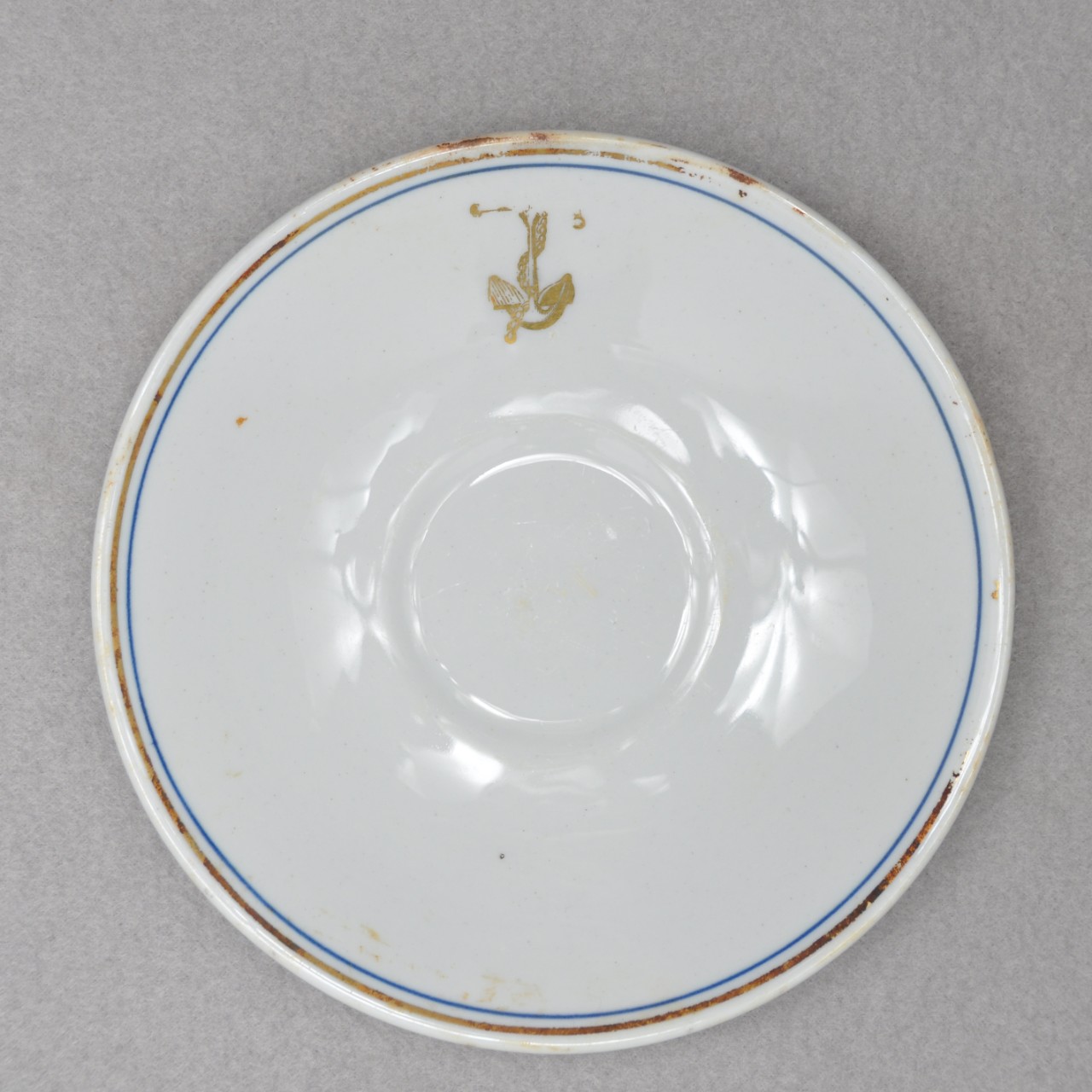 <p>A small, white ceramic saucer with a gold and blue ring around the rim. There is also a gold anchor with a chain wrapped around it located on the lip. On one side of the plate is a small crack.</p>