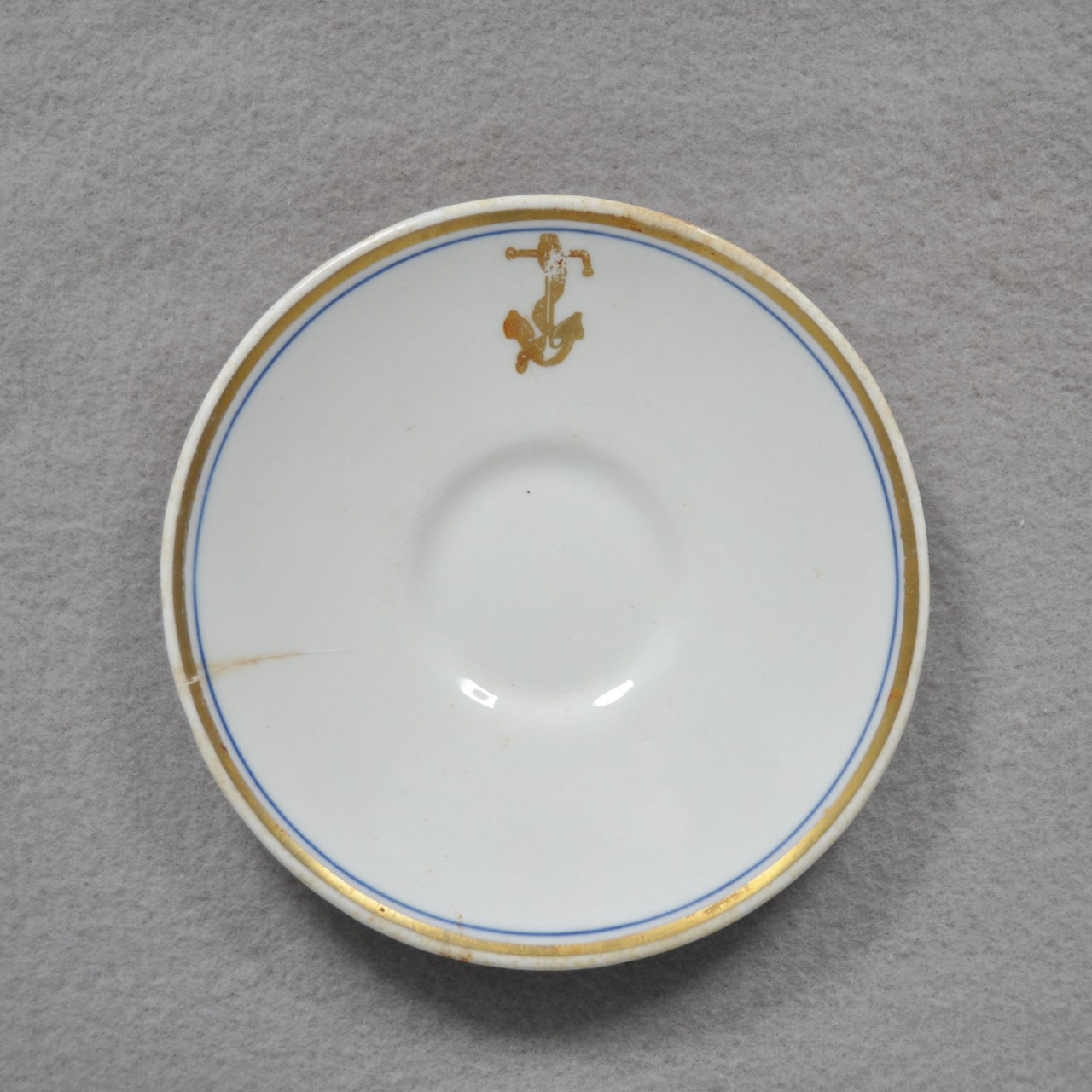 <p>A small, white ceramic saucer with a gold and blue ring around the rim. There is also a gold anchor with a chain wrapped around it located on the lip. On one side of the plate is a small crack.</p>
