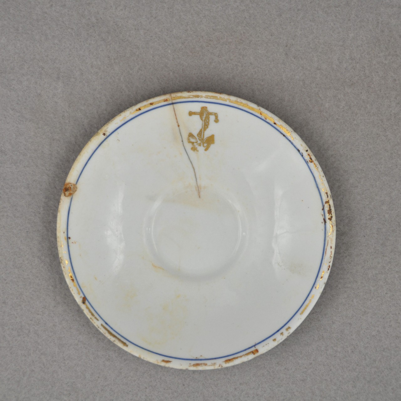 <p>A small white ceramic saucer with a gold and blue ring around the rim. A gold anchor with a chain wrapped around it is located on the lip. Near the anchor is a crack that goes to the middle of the saucer.</p>