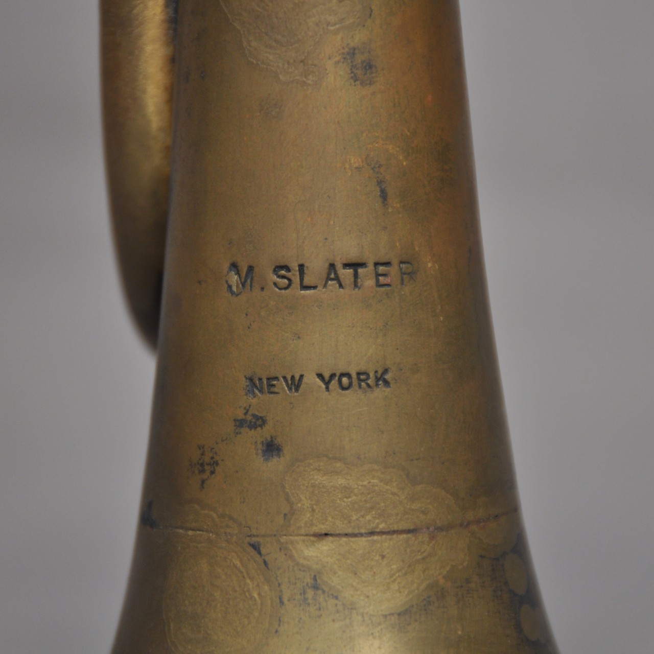 <p>Close-up of a brass bugle’s bell with the marker’s mark stamped on it. It reads, “M. SLATER” on top with “NEW YORK” below it.</p>
