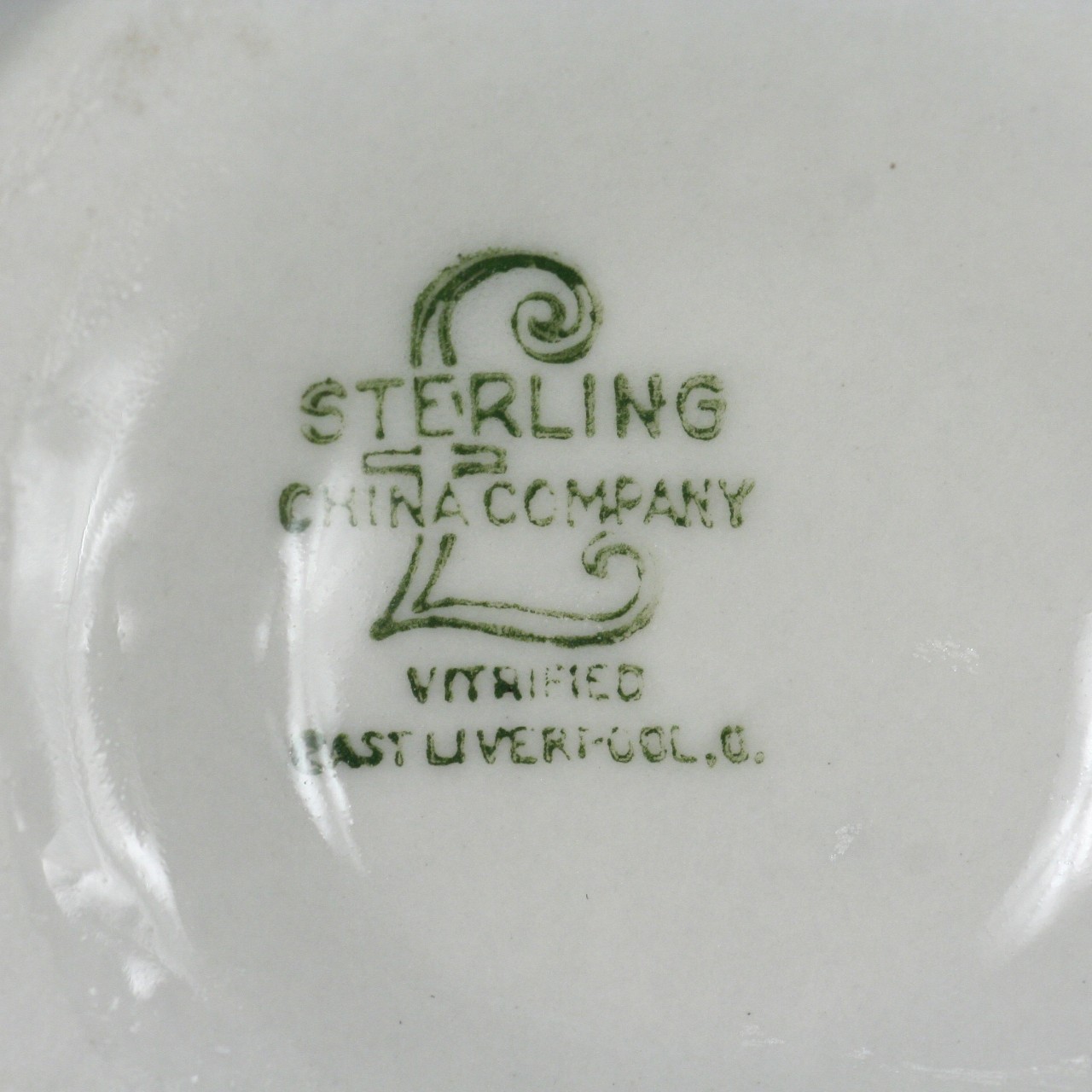 <p>A close up of the maker’s mark located on the base of a white cup. It has a large L with “Sterling China Company” across the L. Below the large L is the words “Vitrified East Liverpool”.</p>
