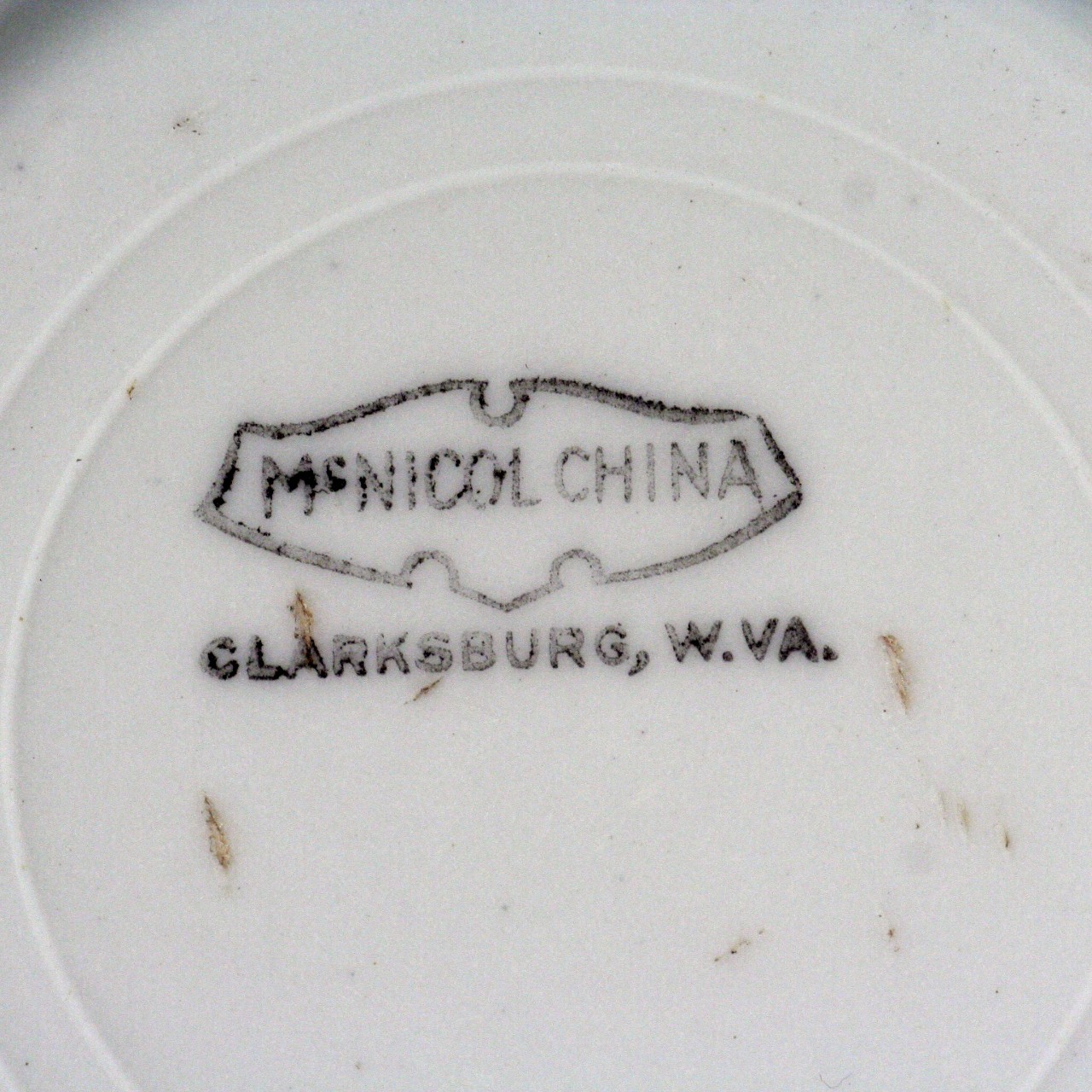 <p>Close up of the cup's maker’s mark. It is “McNicol China” surrounded by a simple black line motif. Underneath the motif is the words “Clarksburg. W. Va.”.</p>
