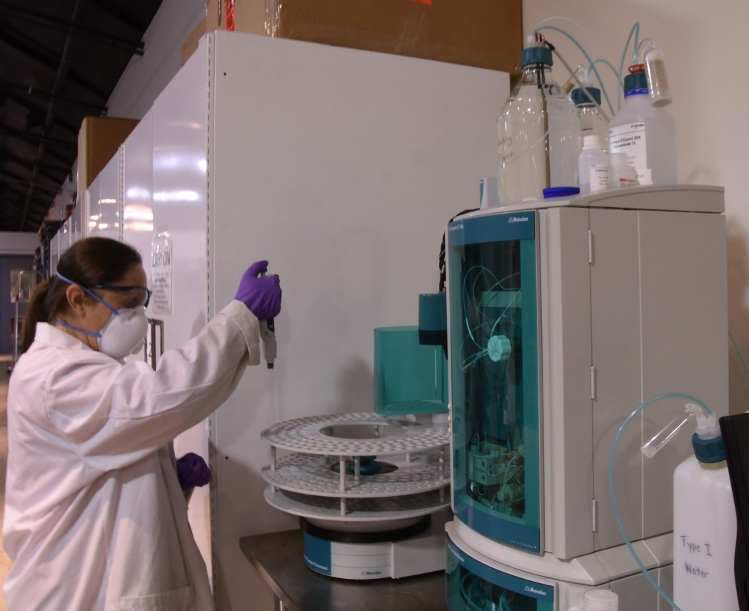 Color photo of a woman wearing a lab coat, mask, protective goggles, and purple gloves putting solution from a pipette into a clear tube held by an ion chromatography system resting on a steel table. 