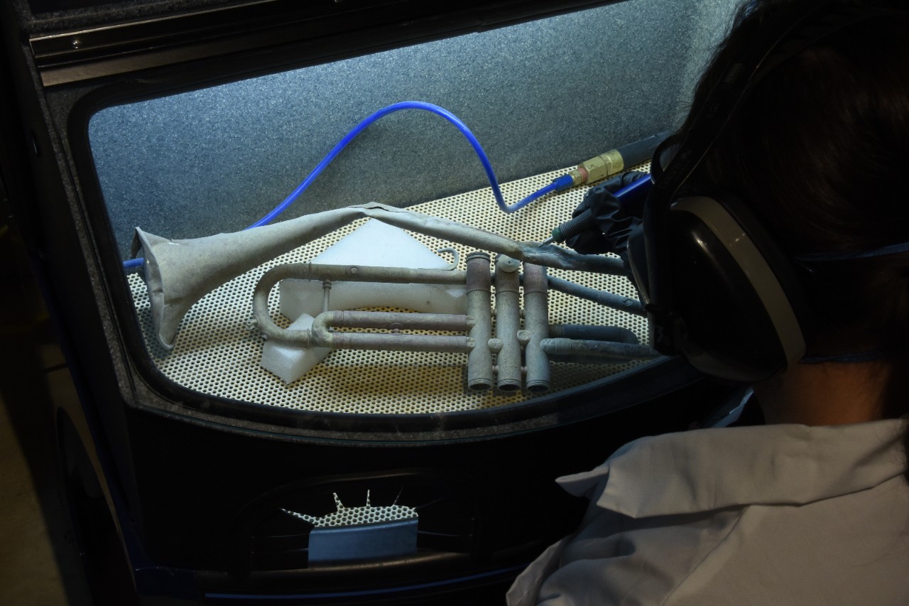 Color photo of a bent trumpet enclosed in a box with a clear glass window. A woman in a lab coat and hearing protection holds an instrument to clean the trumpet's surface through a hole in the side of the box.