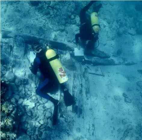 Two scuba divers with clipboards hover over exposed wood on a wreck site in clear blue water. 