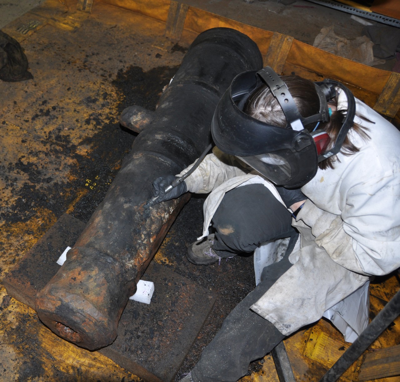 Color photo of a woman in a white lab coat wearing a mask, clear shield, and hearing protection. She is kneeling next to a cannon holding a small pneumatic tool to remove concretion from the cannon's surface.