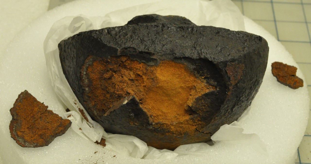 Color photo of a semi-circular iron object resting on white tissue paper. The center shows sections of the iron falling off exposing red corrosion inside.