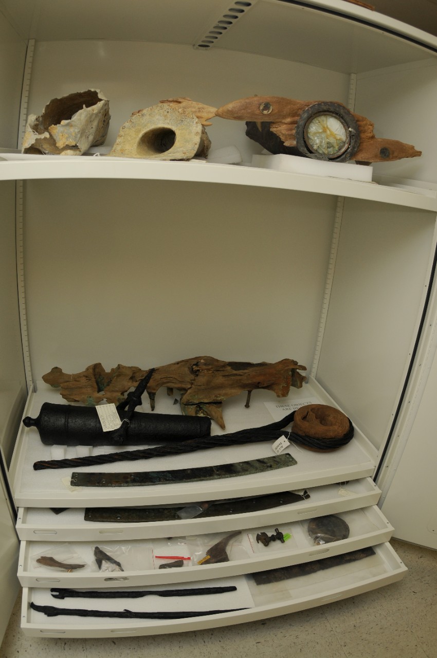Color photo of a white cabinet with two shelves and three pull out drawers. Each shelf and drawer contains archaeological artifacts of various sizes and colors, including a swivel gun and a deadeye..