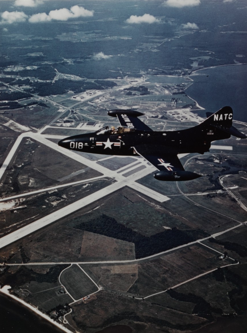 Aerial color photograph of a Grumman F9F-2 Panther with the runways of NAS Patuxen River below it. 