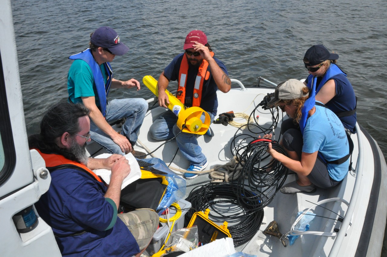 Color photograph of five people sitting on the bow of a small vessel preparing cables and a towfish for deployment.