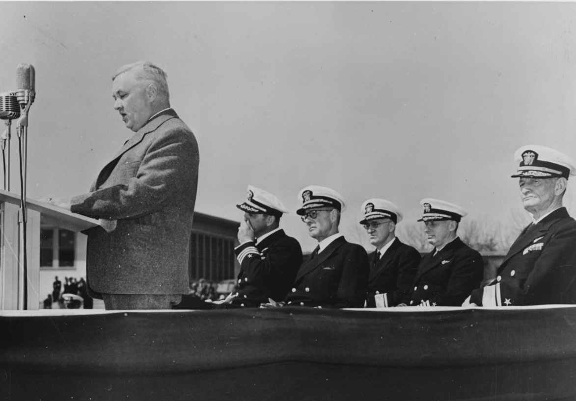 Black and white photograph of a man in civilian clothing standing at a podium (left) with five Navy officers seated behind him.