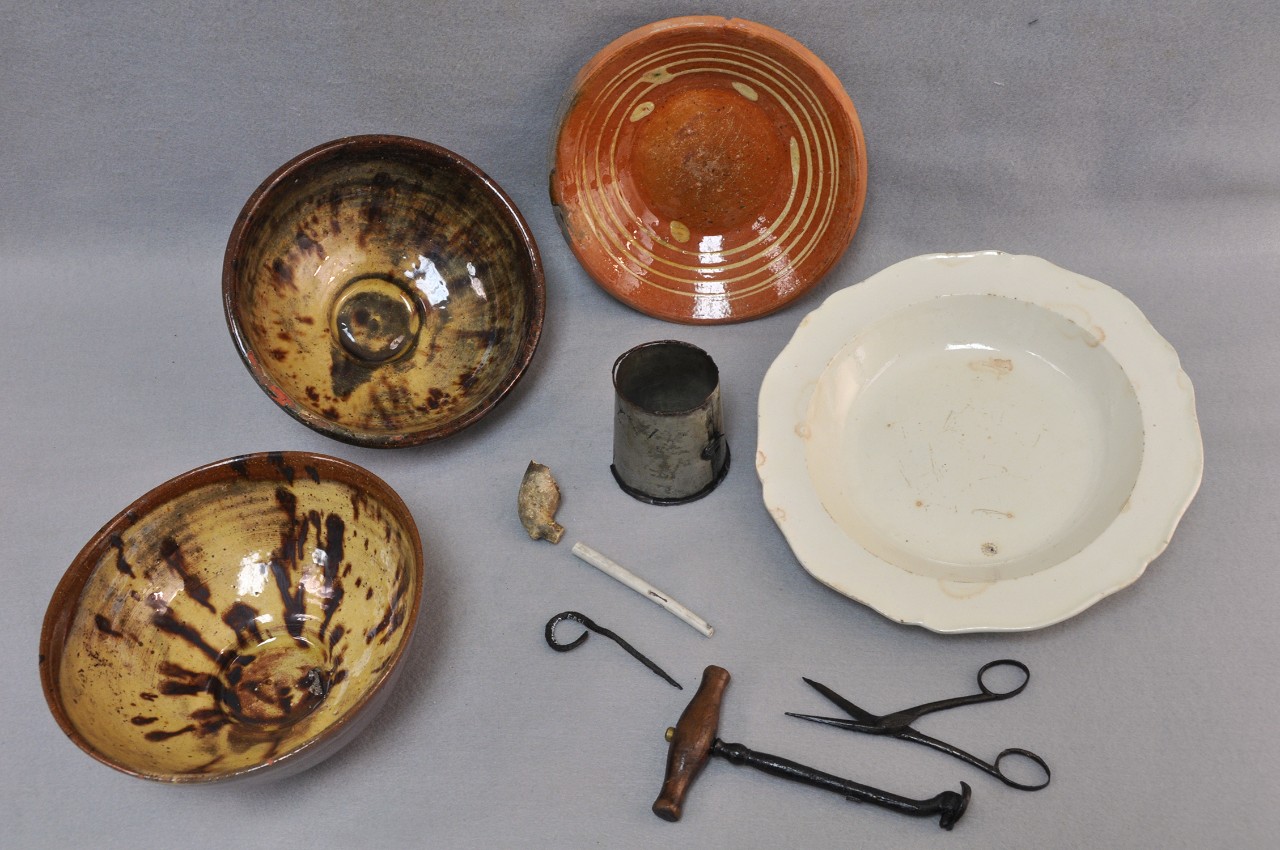 A color photo of three glazed bowls, a white plate, a tin cup, a clay pipe, a iron artifact, a tooth key, and a pair of surgical scissors.