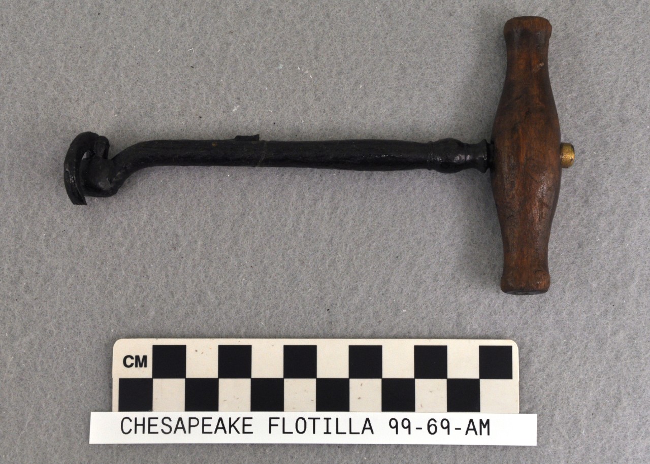 Color photo of an instrument with a wooden handle mounted perpendicular to a slender iron shaft with a hinged hook curving around the tip. The accompanying metric scale indicates it is approximately 15 centimeters long.