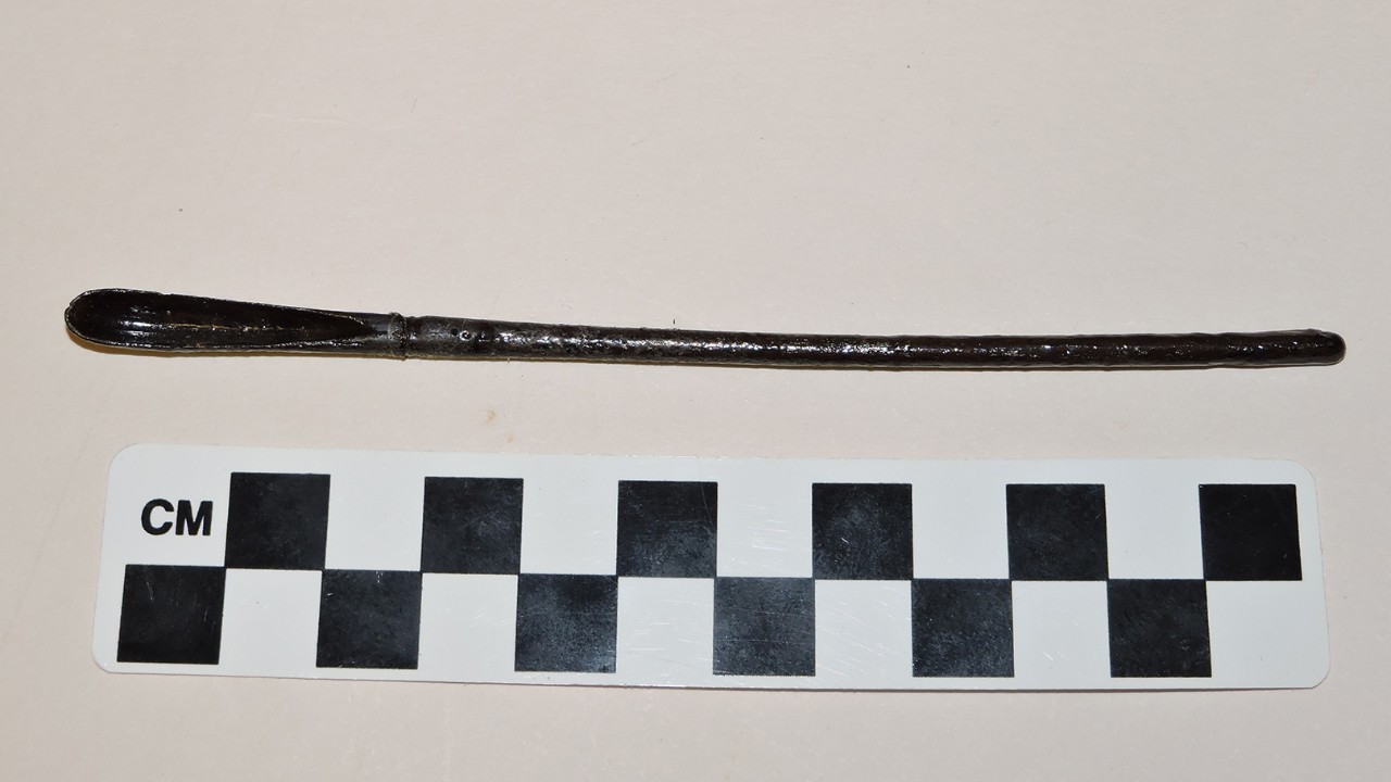 Color photo of a thin iron rod with a groove down one side and a scoop at the end. A metric scale below the object indicates it is just over 12 centimeters long.