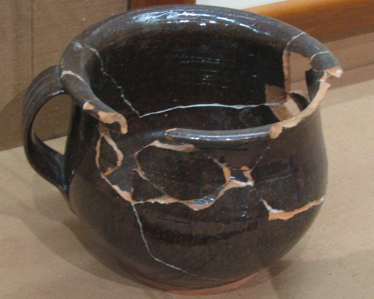 Color photo of a dark brown ceramic pot with a handle and a wide opening. Previously broken, it has been glued back together but several pieces are missing.