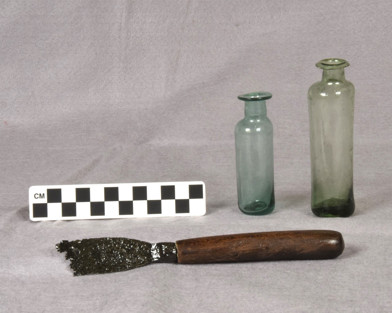 Color image of two small glass bottles with narrow necks and flared rims, and an iron and wood spatula, along with a metric scale that indicates that bottles are approximately 8 and 11 centimeters high.