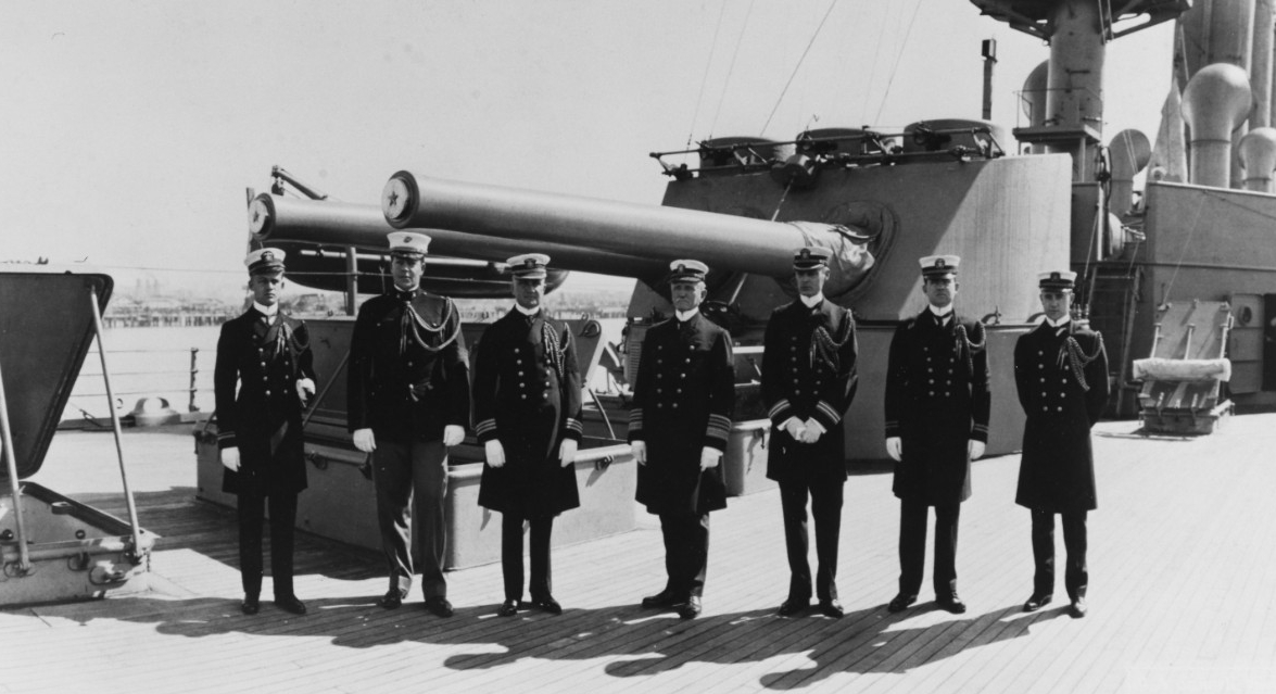 Seven high ranking members of US Navy on the deck of USS San Diego. Behind the men are two large guns attached to the deck.