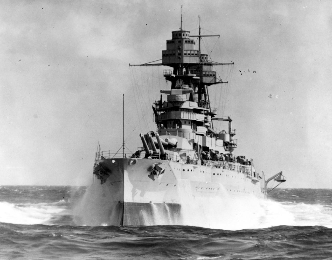 Black and white photo of USS Arizona (BB-39) looking head on at the bow with sea spray breaking off the sides of the ship.